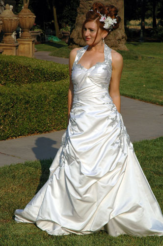 Double faced silk satin halter style gown featuring a twist bodice and rouching to hem. Hand painted silver lace accents with crystal rhinestone.
